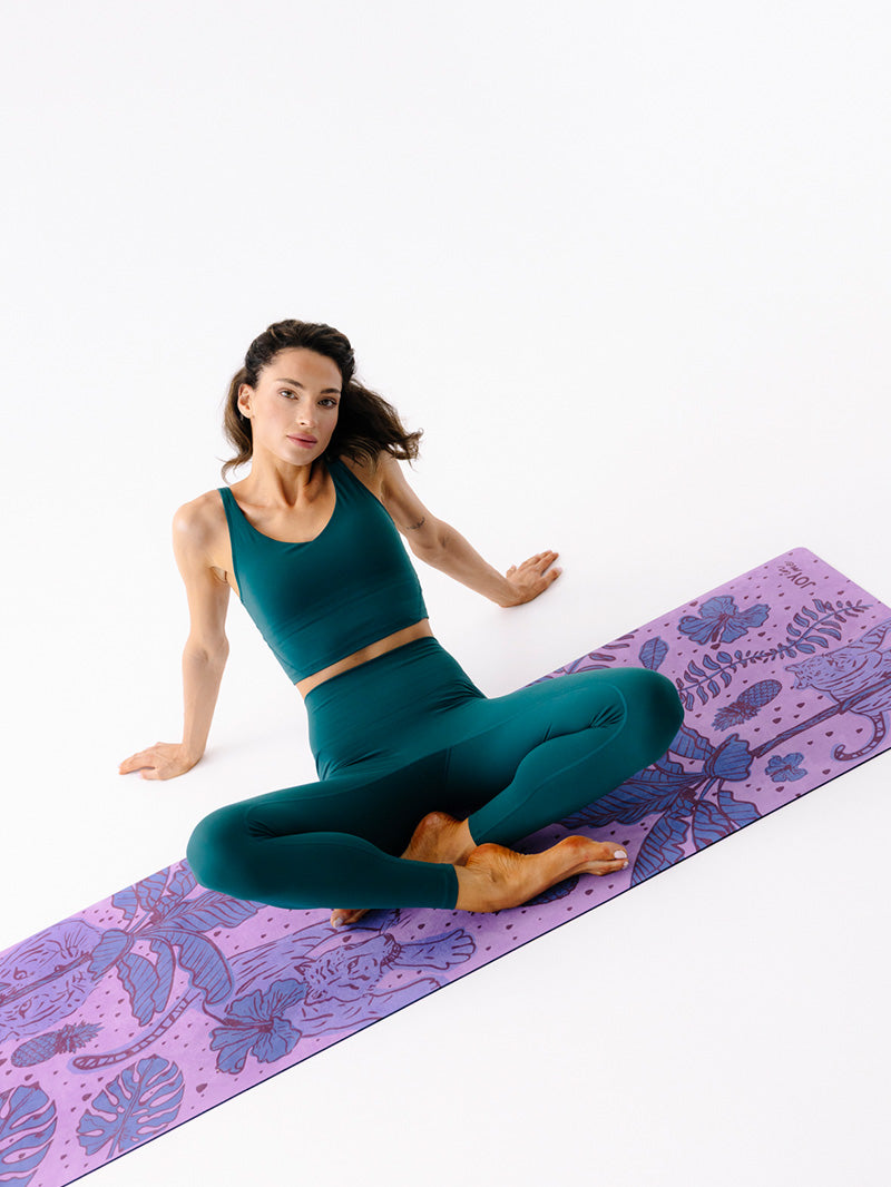 Fioletowa, składana mata do ćwiczeń jogi z kauczuku 1 mm z tropikalnym wzorem - rozłożona mata z siedzącą joginką w zielonym stroju do ćwiczeń